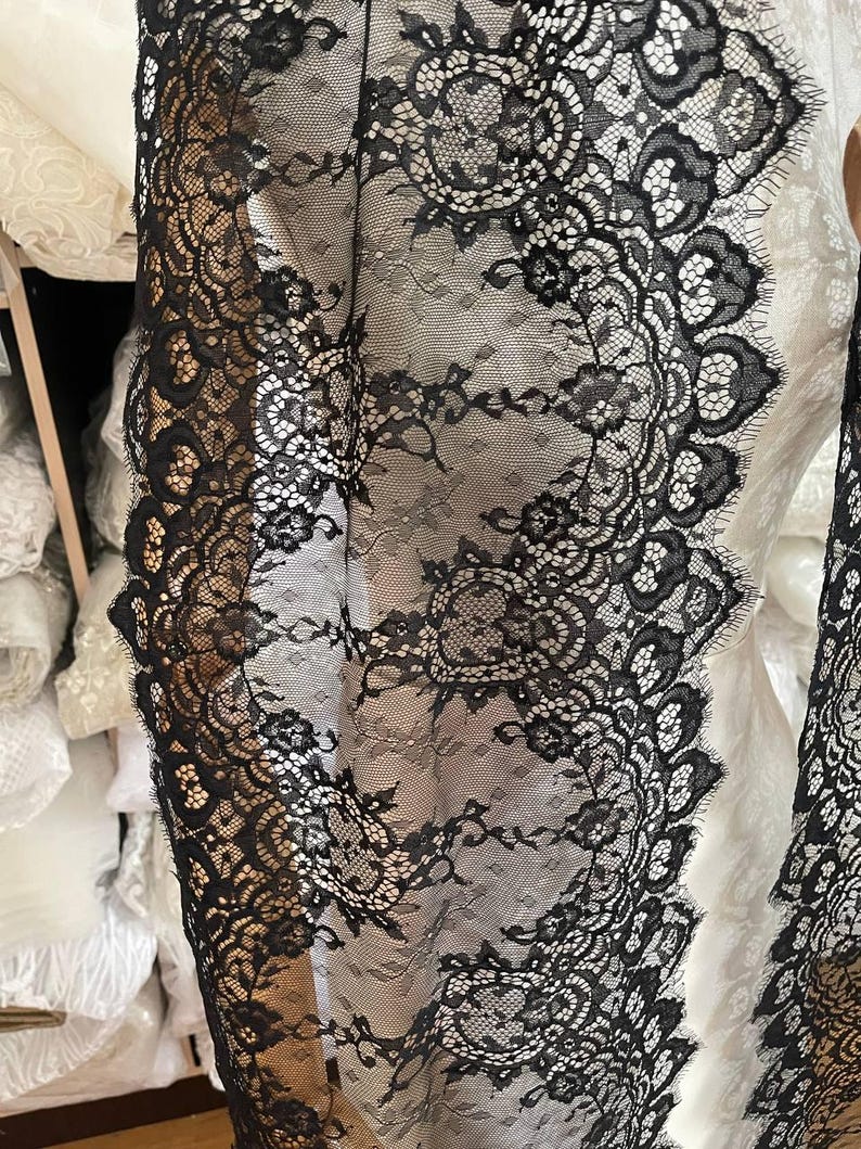 Peut inclure: Gros plan sur un tissu en dentelle noire avec un motif floral. La dentelle a un bord festonn&eacute; et est superpos&eacute;e &agrave; un tissu blanc. Le design complexe et la texture d&eacute;licate sont visibles. Le tissu est probablement utilis&eacute; pour l'habillement ou la d&eacute;coration int&eacute;rieure.