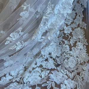 Puede incluir: Primer plano de tela de encaje floral blanco. El delicado encaje presenta flores y hojas bordadas detalladas, creando un dise&ntilde;o texturizado y elegante. La tela es semitransparente, con una ca&iacute;da suave y fluida, perfecta para ropa de novia o formal.