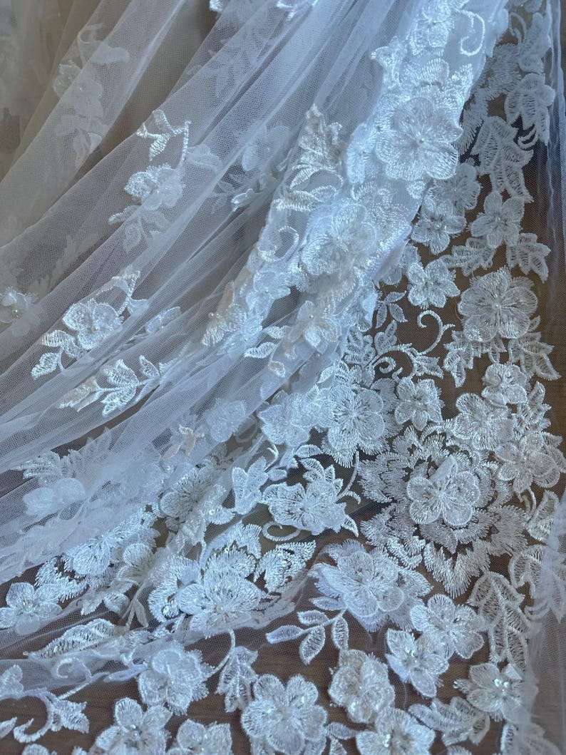 Puede incluir: Primer plano de tela de encaje floral blanco. El delicado encaje presenta flores y hojas bordadas detalladas, creando un dise&ntilde;o texturizado y elegante. La tela es semitransparente, con una ca&iacute;da suave y fluida, perfecta para ropa de novia o formal.