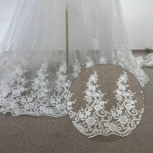 May include: White floral lace fabric with a detailed design. The lace features a repeating pattern of flowers and swirling vines. The fabric is likely used for bridal wear or other formal garments. The image includes a close-up of the lace pattern.