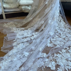 Puede incluir: Un velo de novia blanco con intrincados detalles de encaje floral. La tela transparente y fluida presenta delicadas aplicaciones de flores tridimensionales. Este elegante velo es ideal para bodas, ofreciendo un aspecto rom&aacute;ntico y sofisticado.