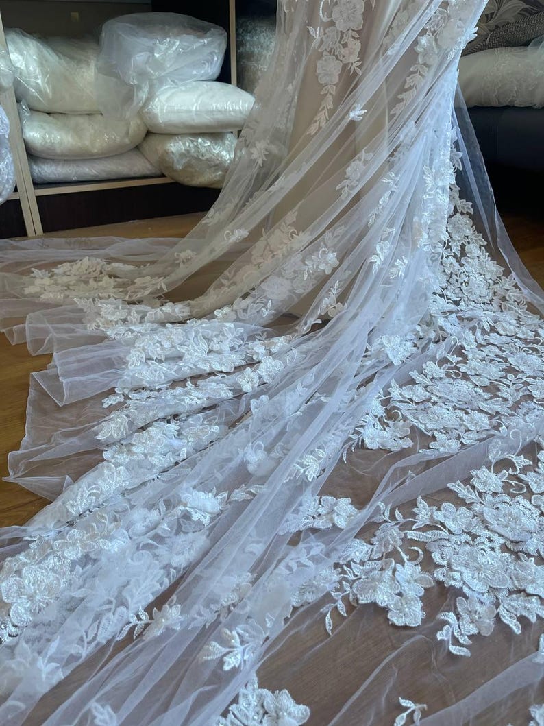 Puede incluir: Un velo de novia blanco con intrincados detalles de encaje floral. La tela transparente y fluida presenta delicadas aplicaciones de flores tridimensionales. Este elegante velo es ideal para bodas, ofreciendo un aspecto rom&aacute;ntico y sofisticado.