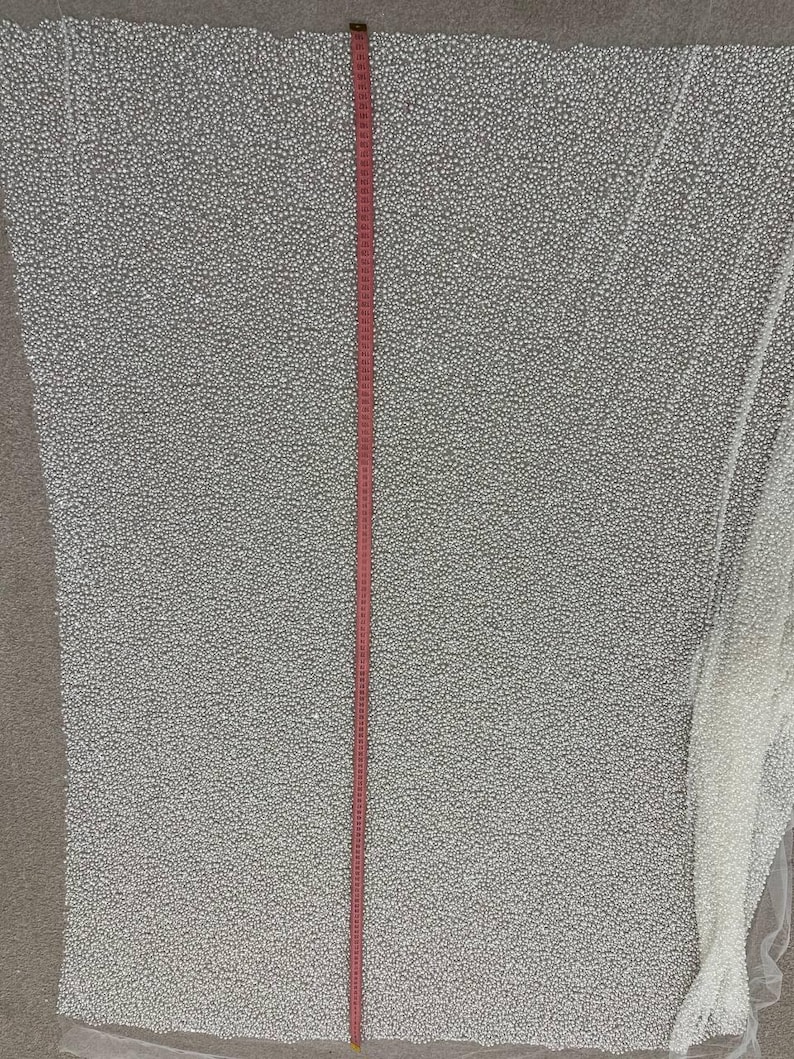 May include: A large piece of white fabric with a dense pattern of small, raised dots. A red measuring tape runs vertically down the center, indicating length. The fabric appears to be a decorative textile, possibly for apparel or crafts.