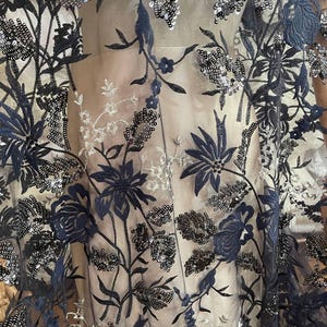 Puede incluir: Tejido transparente con bordado floral azul marino y lentejuelas plateadas. El diseño presenta flores, hojas y tallos detallados. El tejido es semitransparente, revelando un fondo neutro.