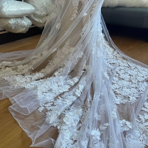 Puede incluir: Un velo de novia blanco con intrincados detalles de encaje floral. La delicada tela cae con gracia, mostrando el bordado detallado de flores y hojas. Un accesorio nupcial.