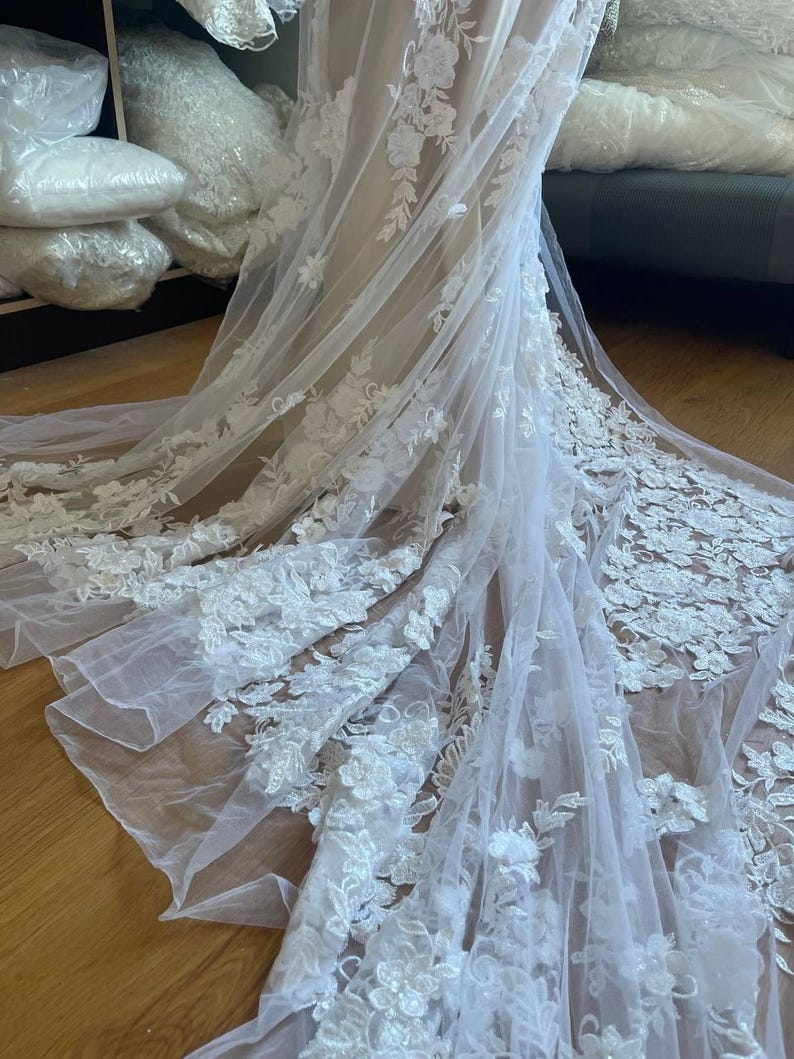 Puede incluir: Un velo de novia blanco con intrincados detalles de encaje floral. La delicada tela cae con gracia, mostrando el bordado detallado de flores y hojas. Un accesorio nupcial.