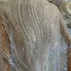 May include: A sheer, white fabric with intricate bead and sequin embellishments. The design features vertical, flowing patterns of sequins and beads, creating a shimmering effect. The fabric is draped over a mannequin, showcasing its texture and drape.