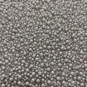 May include: Close-up of a fabric covered in white pearls, sequins, and small beads. The pearls vary in size and are densely packed, creating a textured surface. The background is a light gray color.
