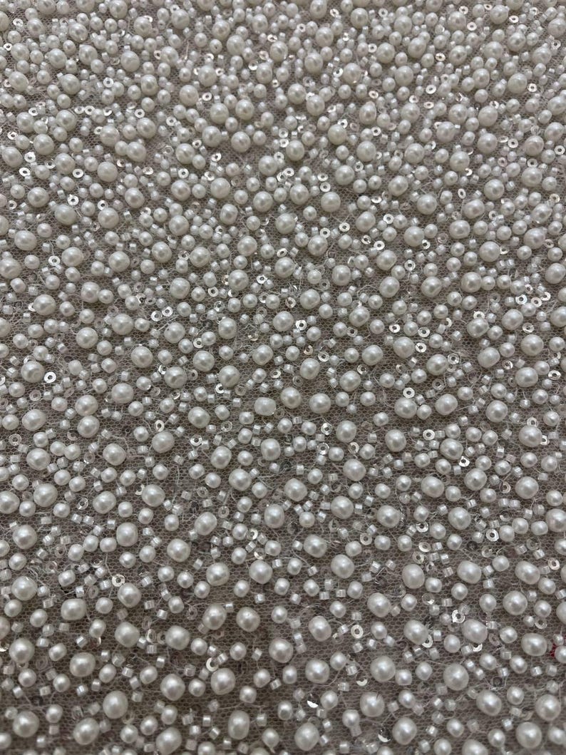 May include: Close-up of a fabric covered in white pearls, sequins, and small beads. The pearls vary in size and are densely packed, creating a textured surface. The background is a light gray color.