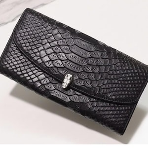 Peut inclure: Un portefeuille pochette noir avec un motif texturé semblable à de la peau de serpent. Le portefeuille est doté d'un fermoir argenté en forme de tête de serpent. La fermeture à rabat est incurvée.