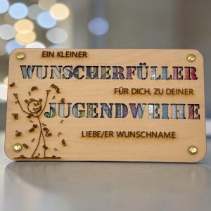 May include: A wooden plaque with the words "EIN KLEINER WUNSCHERF&Uuml;LLER F&Uuml;R DICH, ZU DEINER JUGENDWEIHE" and "LIEBE/ER WUNSCHNAME" engraved. The letters are filled with colorful designs. A stick figure is drawn on the left.