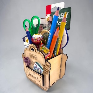May include: A wooden pencil holder shaped like a backpack, filled with pencils, scissors, and candy. The holder has the text "1 Klasse" and a name tag that says "Jannes". The pencils are blue, orange, and yellow.