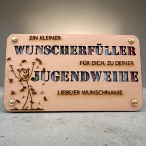 May include: A wooden plaque with cut-out lettering and a stick figure celebrating with money. The text reads "EIN KLEINER WUNSCHERF&Uuml;LLER F&Uuml;R DICH, ZU DEINER JUGENDWEIHE LIEBE/ER WUNSCHNAME". Gold-colored rivets secure the corners.