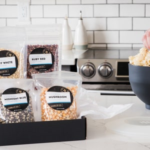 Midnight blue, mushroom, ruby red and baby white popcorn kernel bags in open black gift box on kitchen counter next to large black popcorn bowl. Dell Cove Spices