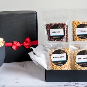 Ruby red, movie theater, mushroom and baby white popcorn kernel bags in open black gift box next to black gift box tied closed with red ribbon and large bowl of popcorn. Dell Cove Spices
