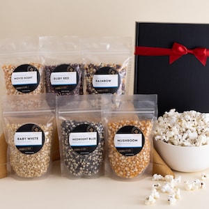 Baby white, midnight blue, mushroom, movie theater, ruby red and rainbow popcorn kernel bags with a black gift box with red ribbon and large white bowl filled with popcorn. Dell Cove Spices