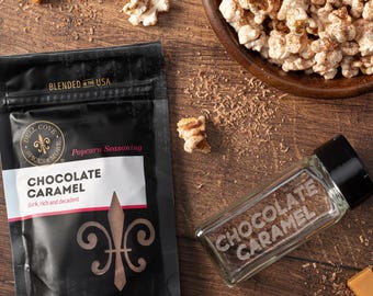 Op de afbeelding: Een houten kom gevuld met chocolade-karamel popcorn, een zwarte zak met het label Dell Cove Spices & More Chocolate Caramel, en een glazen kruidenpotje met een zwarte dop met het label Chocolate Caramel, omringd door karamelblokjes en chocoladeschaafsel.