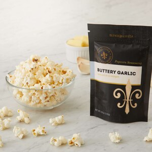 Buttery Garlic seasoning pouch on white counter next to glass bowl of popped popcorn with small white bowl of butter and garlic cloves in the background. Dell Cove Spices