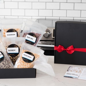 Bags of all 6 kernels types shown in open gift box on kitchen counter next to black gift box with red ribbon and how to make perfect popcorn instruction card. Dell Cove Spices