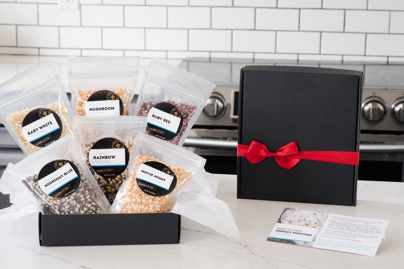 Bags of all 6 kernels types shown in open gift box on kitchen counter next to black gift box with red ribbon and how to make perfect popcorn instruction card. Dell Cove Spices