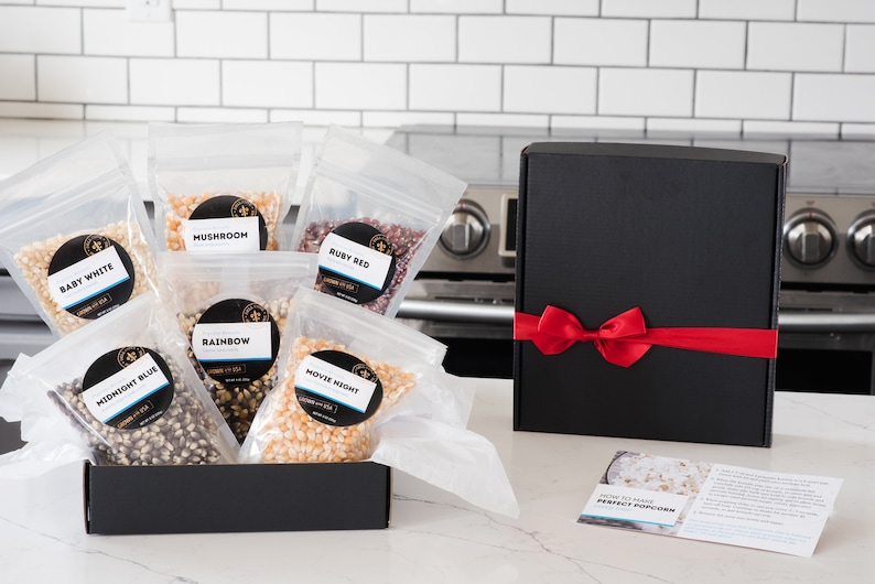 Bags of all 6 kernels types shown in open gift box on kitchen counter next to black gift box with red ribbon and how to make perfect popcorn instruction card. Dell Cove Spices