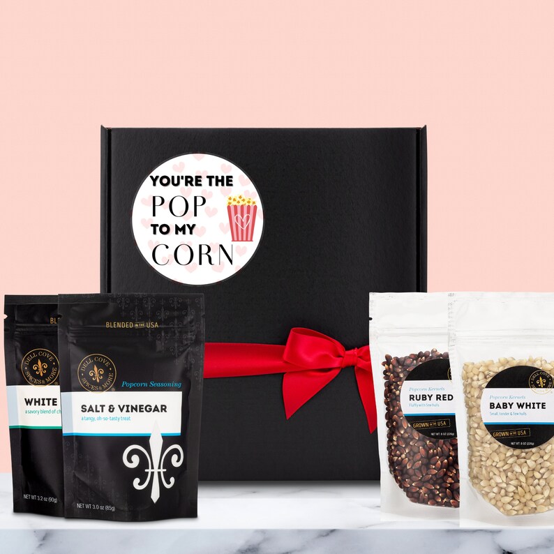 Black gift box with round label that reads you're the pop to my corn with an image of a popcorn box and light pink hearts in the background. The gift box is next to two popcorn seasonings pouches and two popcorn kernel packs. Dell Cove Spices