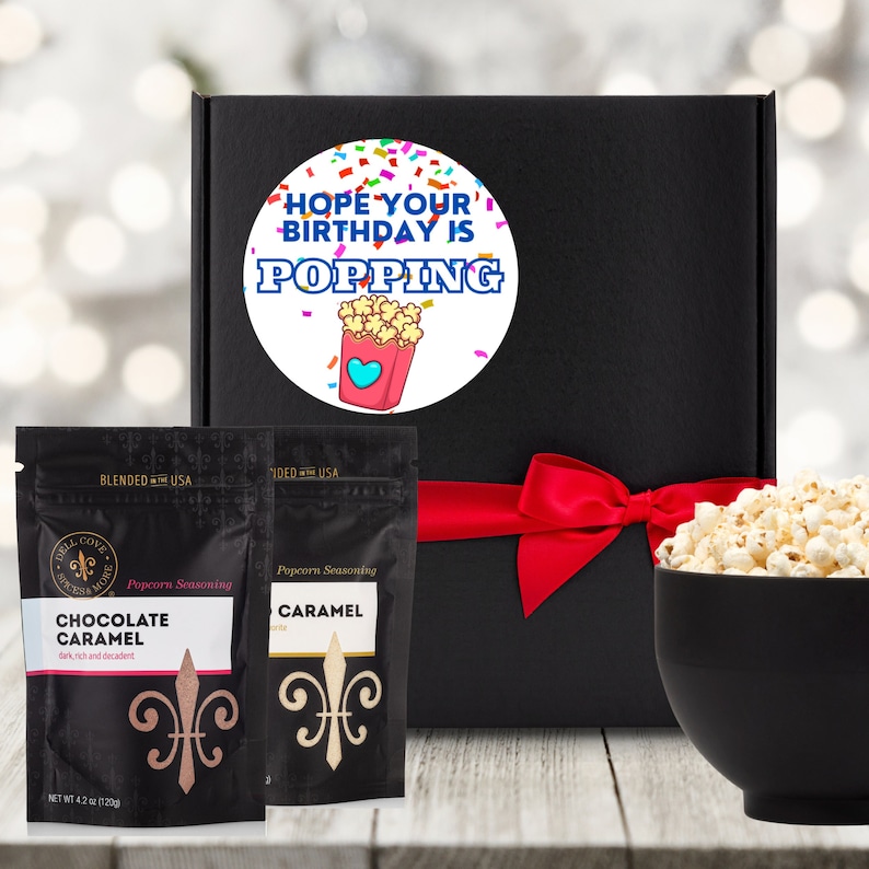 Black gift box tied with red ribbon. Box has round white label that says hope your birthday is popping with confetti and an image of popcorn. A large bowl of popcorn and two seasoning pouches sit in front of gift box. Dell Cove Spices