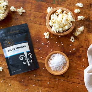 Salt and vinegar seasoning pouch laying on table next to small bowl of salt and two small bowls of popped popcorn. Dell Cove Spices