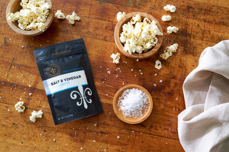 Salt and vinegar seasoning pouch laying on table next to small bowl of salt and two small bowls of popped popcorn. Dell Cove Spices