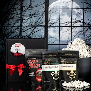 May include: A black gift box with a red bow and a sticker that says "Scary Movie Popcorn Kit". The box contains three bags of popcorn seasoning: White Cheddar, Buttery Garlic, and Scary Movie Popcorn. The box is in front of a window with a full moon and trees.
