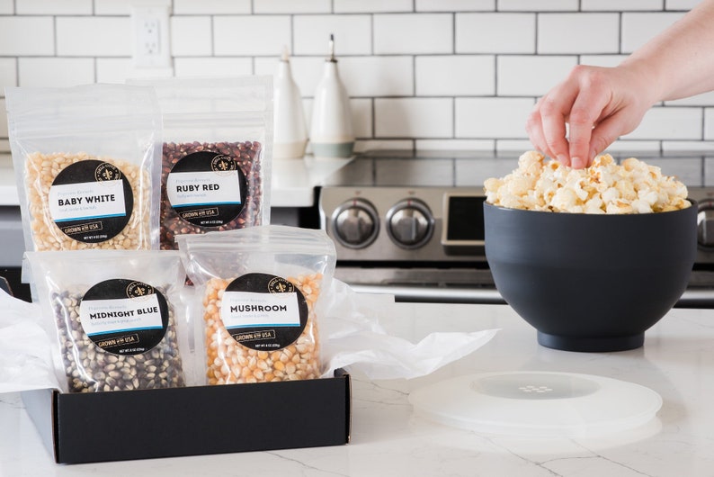 Midnight blue, mushroom, ruby red and baby white popcorn kernel bags in open black gift box on kitchen counter next to large black popcorn bowl. Dell Cove Spices