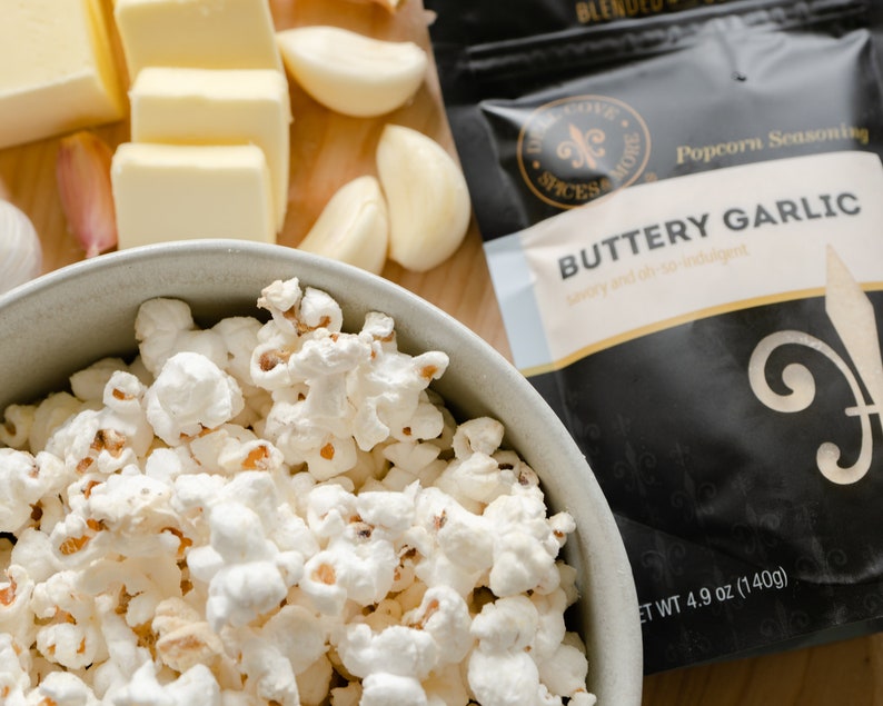 Buttery garlic seasoning pouch next to bowl of popped popcorn, butter and garlic cloves. Dell Cove Spices