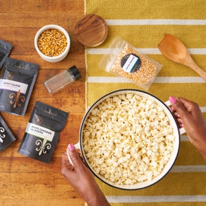 Overhead image of a large kettle filled with popped popcorn on table next to popcorn seasoning pouches, a pack of popcorn kernels, a small bowl filled with kernels and an empty glass seasoning jar. Dell Cove Spices