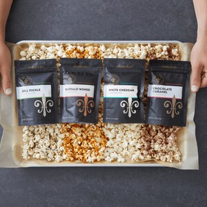 Hands holding out baking tray that has popped seasoned popcorn on it with 4 bags of popcorn seasoning on top of popcorn. Dell Cove Spices