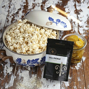 Dill Pickle seasoning pouch on rustic styled table next to jar of pickle slices and a white and blue floral kettle filled with popped popcorn. Dell Cove Spices