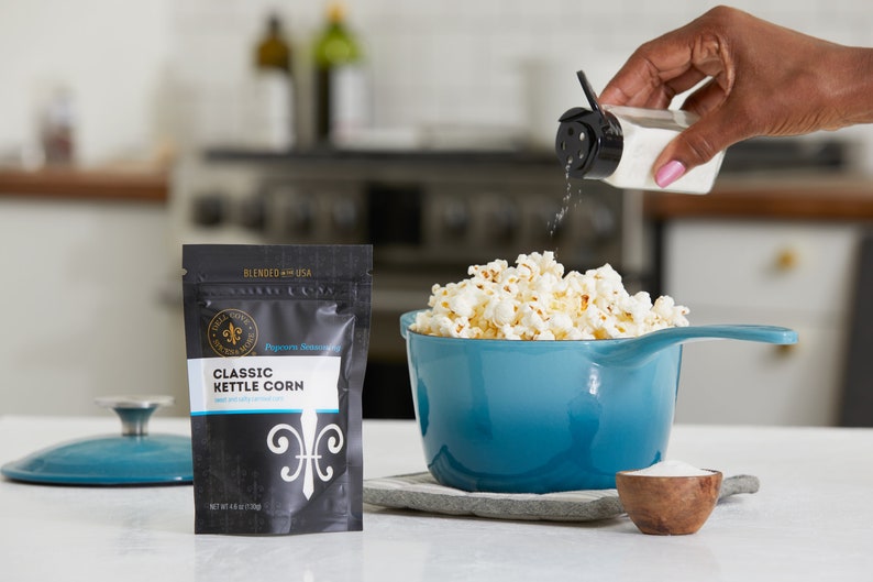 Hand holding glass shaker jar sprinkling seasoning on to popcorn in blue kettle. Classic kettle corn seasoning pouch and a small bowl of seasoning next to kettle on kitchen counter. Dell Cove Spices