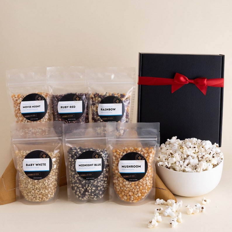 Baby white, midnight blue, mushroom, movie theater, ruby red and rainbow popcorn kernel bags with a black gift box with red ribbon and large white bowl filled with popcorn. Dell Cove Spices
