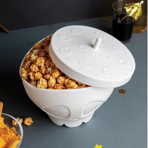 May include: White popcorn bowl with a matching lid featuring skull designs. The bowl is filled with caramel popcorn. A glass of dark liquid with a cocktail umbrella is in the background. A fun and festive serving dish.