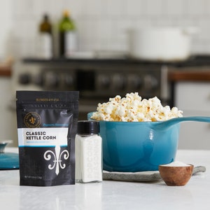 Blue kettle on counter next to classic kettle corn seasoning pouch, small wooden bowl filled with seasoning and glass shaker filled with seasoning. Dell Cove Spices