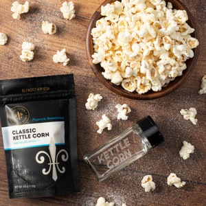 Puede incluir: Vista cenital de palomitas de ma&iacute;z, un cuenco de madera lleno de palomitas y un agitador de vidrio con la inscripci&oacute;n "Kettle Corn". Tambi&eacute;n hay una bolsa negra de condimento para palomitas. Las palomitas est&aacute;n esparcidas sobre una superficie de madera.