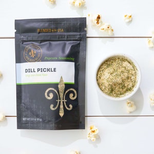 Dill Pickle seasoning pouch laying on white table with small bowl of seasoning and popped popcorn sprinkled around. Dell Cove Spices