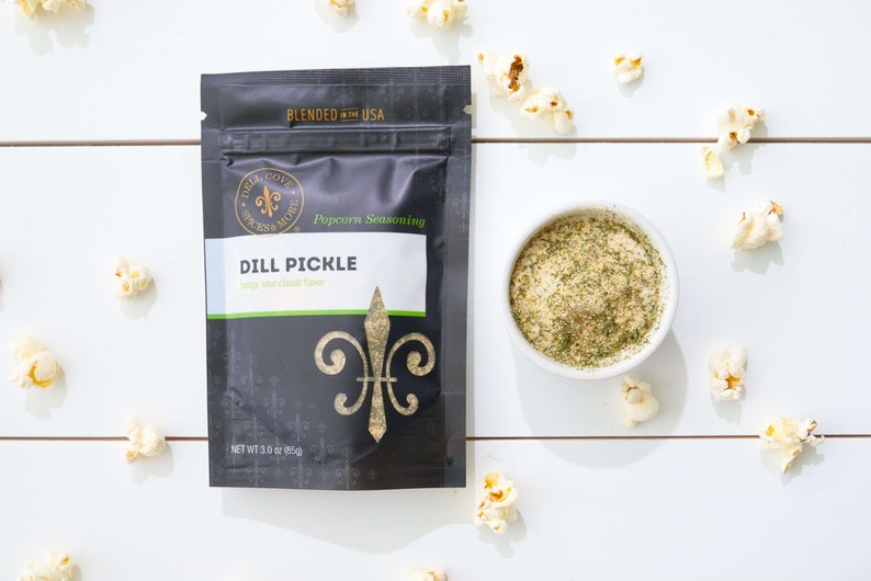 Dill Pickle seasoning pouch laying on white table with small bowl of seasoning and popped popcorn sprinkled around. Dell Cove Spices