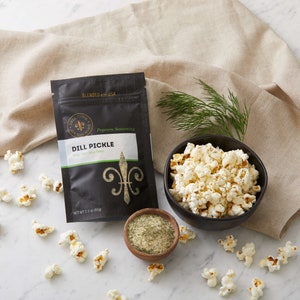 Dill pickle seasoning pouch on marble counter with small bowl of popped popcorn, small bow of seasoning and fresh dill. Dell Cove Spices