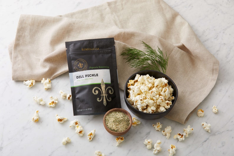 Dill pickle seasoning pouch on marble counter with small bowl of popped popcorn, small bow of seasoning and fresh dill. Dell Cove Spices