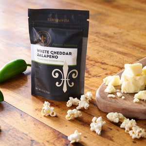 White cheddar jalapeno seasoning pouch on table next to cutting board with white cheddar cheese, whole jalapenos and popped popcorn sprinkled around. Dell Cove Spices