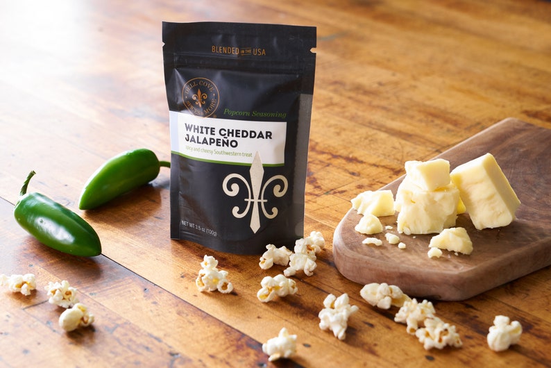 White cheddar jalapeno seasoning pouch on table next to cutting board with white cheddar cheese, whole jalapenos and popped popcorn sprinkled around. Dell Cove Spices