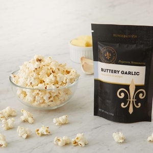 Buttery garlic seasoning pouch on counter with small glass bowl of popped popcorn, a small dish of butter and garlic in the background and popped popcorn sprinkled around. Dell Cove Spices