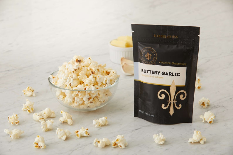 Buttery garlic seasoning pouch on counter with small glass bowl of popped popcorn, a small dish of butter and garlic in the background and popped popcorn sprinkled around. Dell Cove Spices