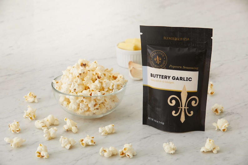 Buttery Garlic seasoning pouch on white counter next to glass bowl of popped popcorn with small white bowl of butter and garlic cloves in the background. Dell Cove Spices