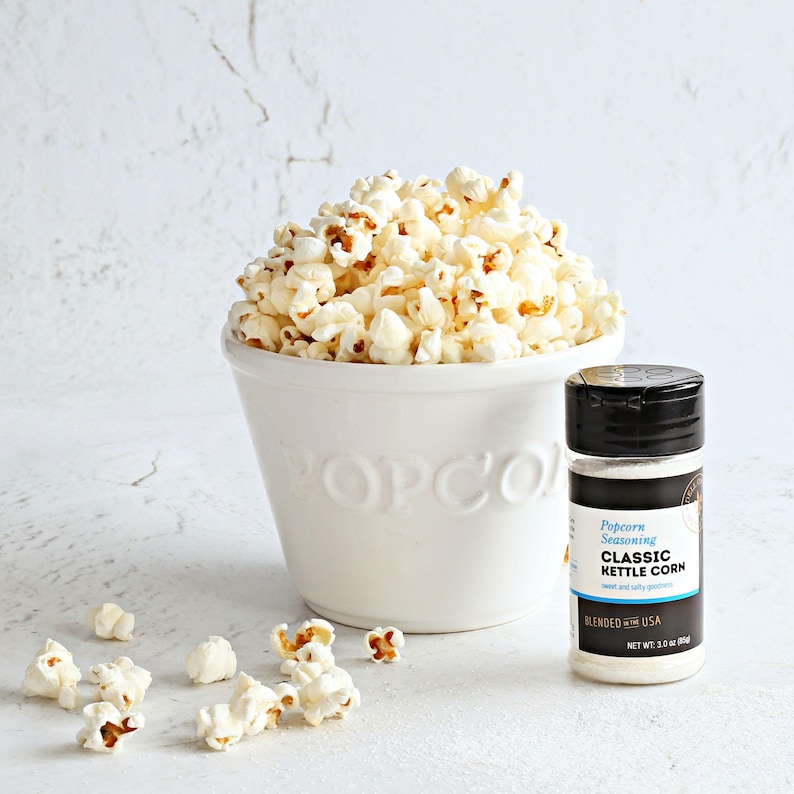 Puede incluir: Un cuenco de cer&aacute;mica blanco lleno de palomitas de ma&iacute;z reventadas, con una botella de condimento para palomitas al lado. La botella de condimento para palomitas tiene una tapa negra y una etiqueta que dice "Classic Kettle Corn". Hay trozos de palomitas esparcidos delante del cuenco.